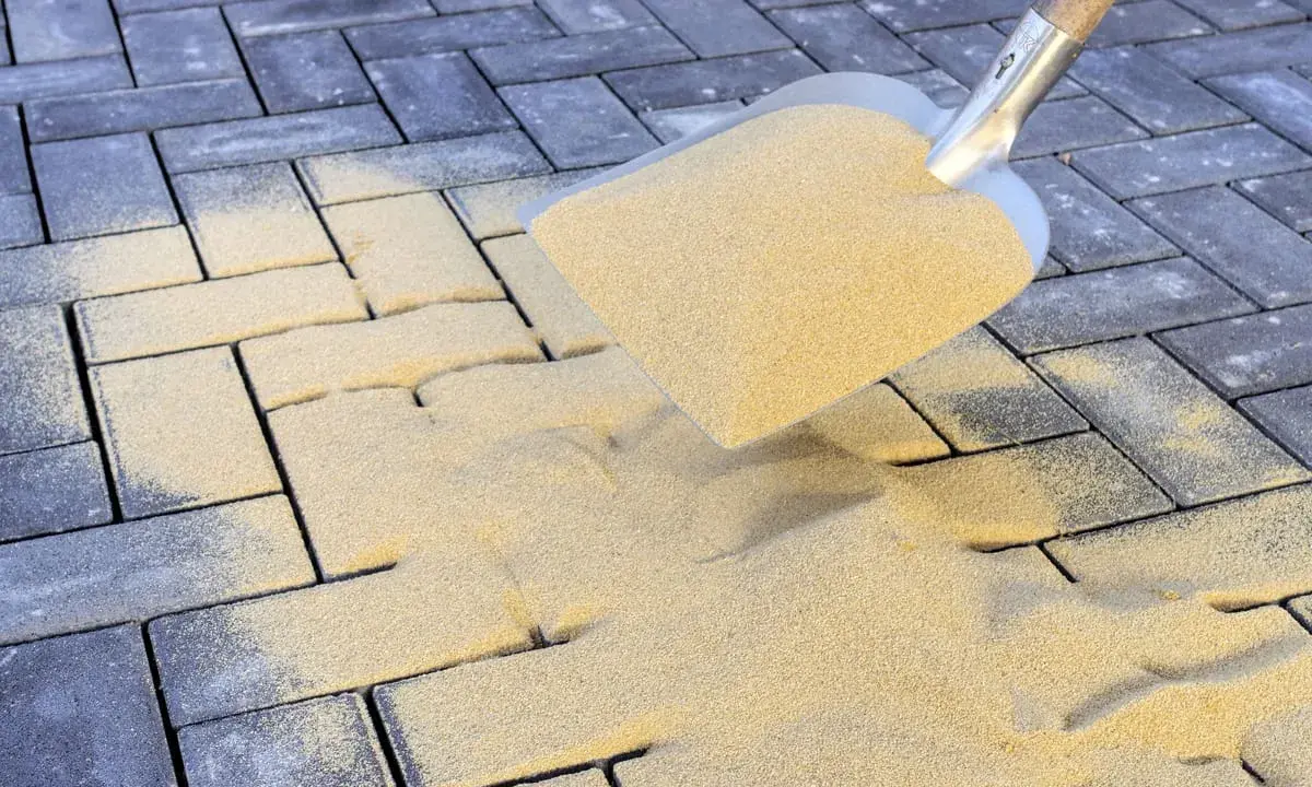 Łopata rozsypuje piasek na kostce brukowej. To kluczowy etap prac, który decyduje, jak długo ma leżeć piasek na kostce brukowej, by zapewnić stabilność.