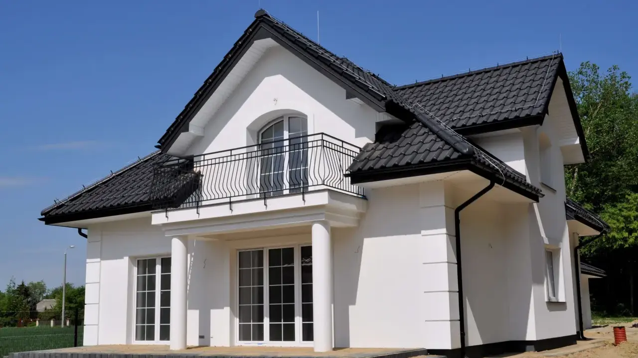 Nowy dom z białym gipsem na zewnątrz, czarnym dachem i balkonem. Wokół plac budowy z piaskiem.