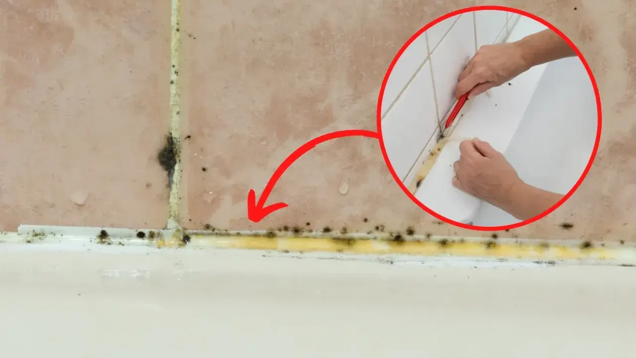 Pokazano, jak usunąć stary silikon z pleśnią. Dłoń z nożykiem usuwa żółty, zapleśniały silikon z fugi przy wannie.