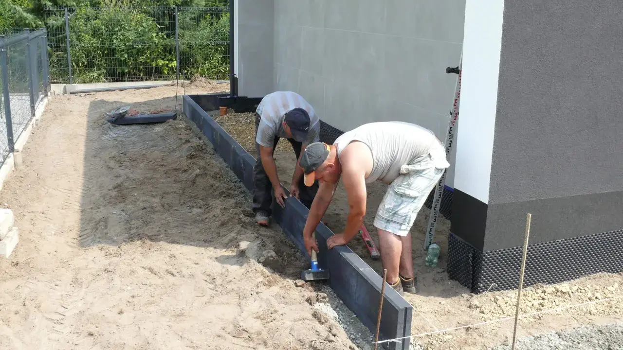 Dwóch mężczyzn zajmuje się układaniem obrzeży betonowych wokół budynku.
