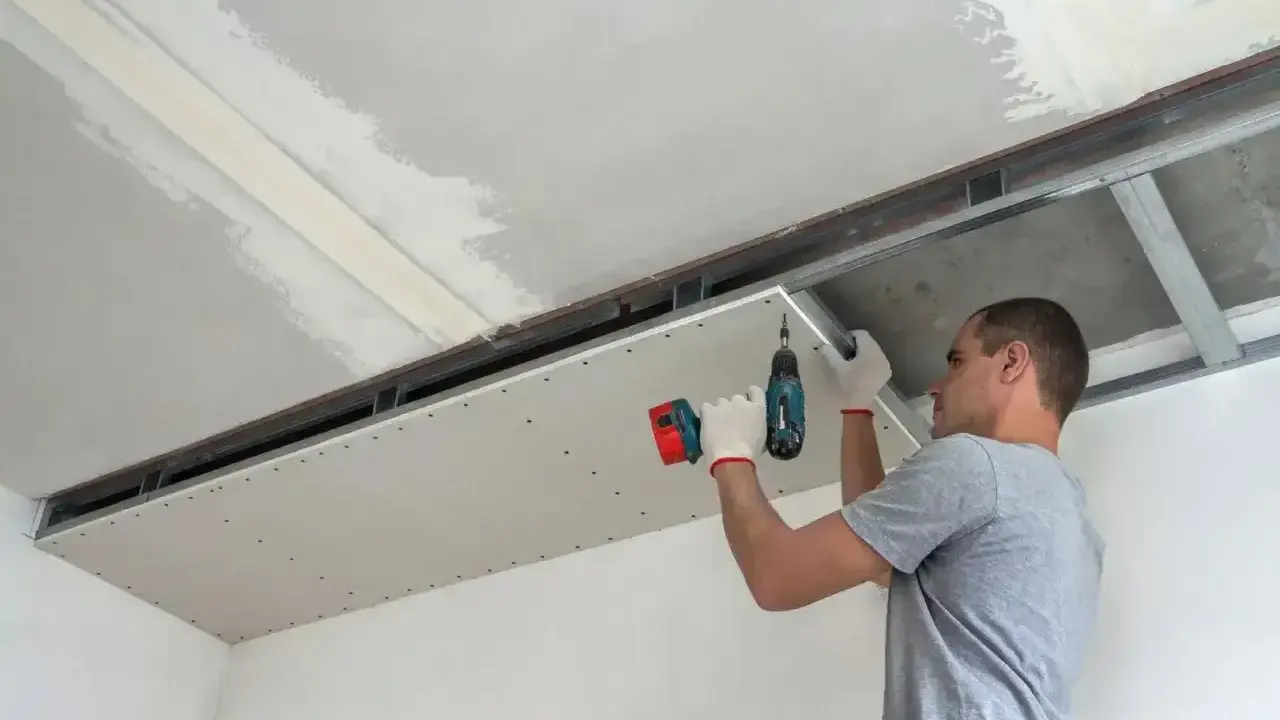 Mężczyzna przykręca płytę kartonowo-gipsową do stelaża sufitowego. Do cięcia karton gips używa się specjalnych narzędzi.