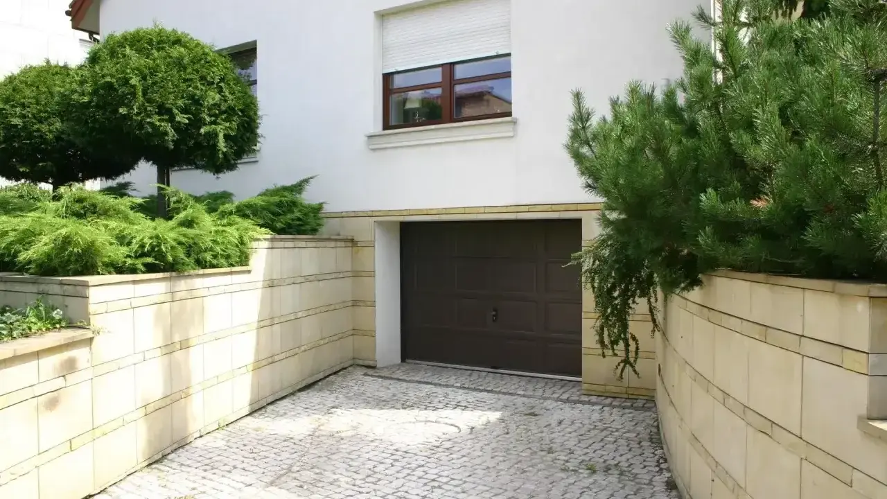 Wjazd z kostki brukowej prowadzi do ciemnobrązowej bramy garażowej. Otaczają go kamienne murki z bujną zielenią.