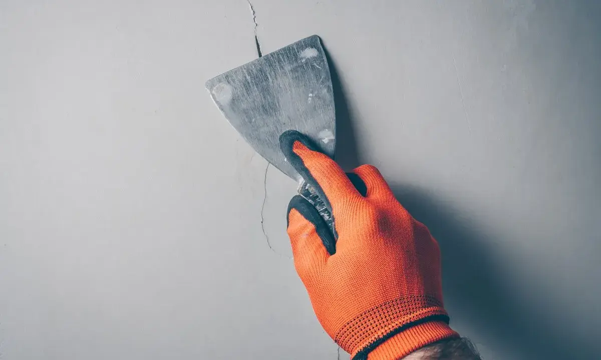 Ręka w pomarańczowej rękawicy zacierająca pionowe pęknięcia ścian działowych metalową szpachlą.