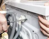 Ręce majsterkowicza przykręcają śrubę przy odpływie pralki bez syfonu. Wąż odpływowy jest podłączony do białego złącza.