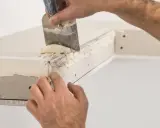 Ręce nakładają masę szpachlową na narożnik zewnętrzny z użyciem kielni. To kluczowy etap, jak szpachlować narożniki zewnętrzne, by uzyskać gładką powierzchnię.