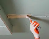 Ręka w rękawicy nakłada masę szpachlową na taśmę zbrojącą, przygotowując do klejenia płyt gk na grzebień.