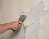 Dłoń zeskrobuje łuszczącą się farbę z gruntem ze ściany.