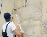 Pracownik w białej koszulce i granatowym ogrodniczkach stosuje agregat do tynkowania stare ściany. Widać przygotowania do prac.