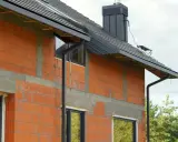 Nowy dom w budowie, z widocznymi cegłami i oknami. Zastanawiasz się, jakie rury do odprowadzenia wody z rynien wybrać? Tutaj widzisz czarne rury spustowe.