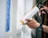 Ręce trzymają pistolet do silikonu, aplikując go na ramę okna. Dowiedz się, jak otworzyć silikon i uszczelnić szczeliny.
