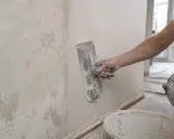 Ręka trzymająca kielnię wygładza ścianę pokrytą starą, łuszczącą się farbą.