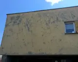 Ściana budynku z żółtym, odpadającym tynkiem. Widać dwa okna z białymi ramami i firankami.
