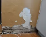 Zła gładź na ścianie, łuszcząca się farba odsłania tynk. Widoczne drewniane belki i biały kabel na podłodze.