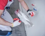 Ręce w rękawiczkach nakładają gips szpachlowy na ścianę za pomocą kielni. Idealny do wygładzania i przygotowania powierzchni.