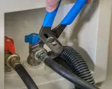 Ręka z niebieskimi szczypcami dokręca zawór do pralki. Obok czerwony zawór i wąż.