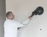 Mężczyzna w okularach ochronnych szlifuje ścianę gładzią z włóknem szklanym, przygotowując ją do malowania.