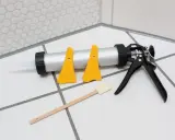 Pistolet do silikonu, żółte szpachelki i drewniana łopatka na płytkach. Czy można kłaść silikon na silikon? Narzędzia do fugowania.