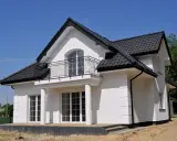 Nowy dom z białym gipsem na zewnątrz, czarnym dachem i balkonem. Wokół plac budowy z piaskiem.