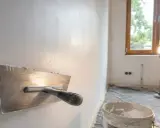 Gips na ścianę jest nakładany kielnią. W tle okno i lampa budowlana.