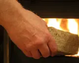Dłoń wkłada do palącego się kominka brykiet z trocin. Dobry wybór, jeśli zastanawiasz się, jaki brykiet do kominka.