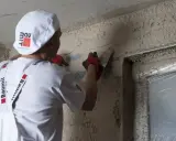 Pracownik w białej czapce i fartuchu wykonuje zacieranie tynku gipsowego na ścianie, używając kielni.