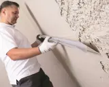 Mężczyzna w białej koszulce i rękawiczkach wygładza ścianę narzędziem. Czy gruntować między warstwami gładzi? To pytanie zadaje sobie wielu.