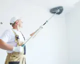 Pracownik w białej czapce i ogrodniczkach czyści sufit specjalną miotłą. Czas schnięcia gruntu przed szpachlowaniem jest kluczowy.