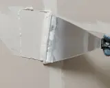 Ręka w rękawicy nakłada masę do łączenia płyt gk na ścianę za pomocą szpachli.