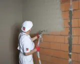 Pracownik w białym stroju nakłada gładź na tynk cementowo wapienny za pomocą agregatu tynkarskiego.