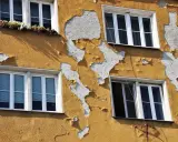Ściana budynku z żółtym tynkiem baranek, który łuszczy się, odsłaniając biały podkład. Widać cztery okna.