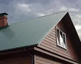Dach domu pokryty zieloną blachą trapezową, z ceglanym kominem i drewnianą ścianą. Precyzyjne cięcie blachy trapezowej zapewnia estetykę.