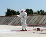 Pracownik w białym kombinezonie natryskuje białą masę na płaski dach, szukając alternatywy dla papy.