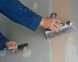 Ręce w niebieskim kombinezonie nakładają gładź szpachlową na ścianę za pomocą kielni. Proces ten przypomina nakładanie gładzi wałkiem, tworząc gładką powierzchnię.