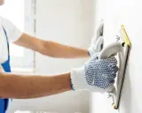 Pracownik w niebieskim ogrodniczkach wygładza ścianę pacą. Zastanawiasz się, ile gładzi na m2 zużyjesz?