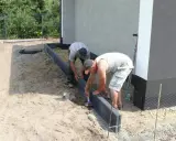 Dwóch mężczyzn zajmuje się układaniem obrzeży betonowych wokół budynku.