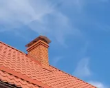 Obróbka komina blachą modułową na dachu z czerwonej dachówki. Niebo błękitne z delikatnymi chmurkami.