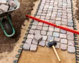 Układanie kostki brukowej z progiem. Wózek z kamieniami, poziomica i gumowy młotek czekają na dokończenie ścieżki.