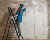 Kobieta na drabinie usuwa gładź ze ściany za pomocą pędzla. Proces przygotowania do remontu.