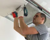 Mężczyzna w białych rękawiczkach przykręca wkrętarką płyty gipsowe do stelaża. Montaż płyt gipsowych na ścianie drewnianej w toku.