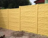 Jasnożółte, betonowe płoty imitujące cegłę, z pionowymi słupkami. Widać fragment podwórka i drzewa w tle.