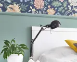Zielona ściana z lamperią i kwiatową tapetą. Obok łóżka stoi lampka i doniczka z rośliną.