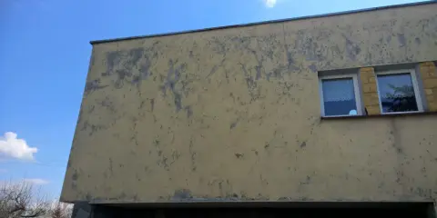 Ściana budynku z żółtym, odpadającym tynkiem. Widać dwa okna z białymi ramami i firankami.