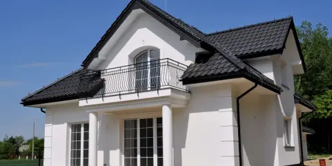 Nowy dom z białym gipsem na zewnątrz, czarnym dachem i balkonem. Wokół plac budowy z piaskiem.