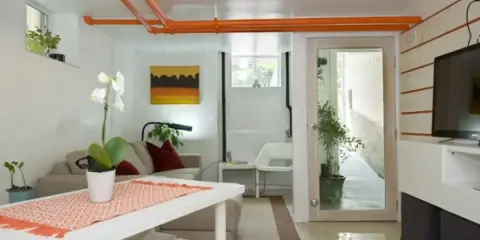 Pomarańczowa zabudowa rur pod sufitem w salonie. Wygodna kanapa, stół z ozdobnym bieżnikiem i telewizor.