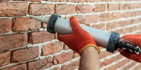 Ręce w pomarańczowych rękawicach używają pistoletu z masą uszczelniającą, by wypełnić ubytki w cegle.