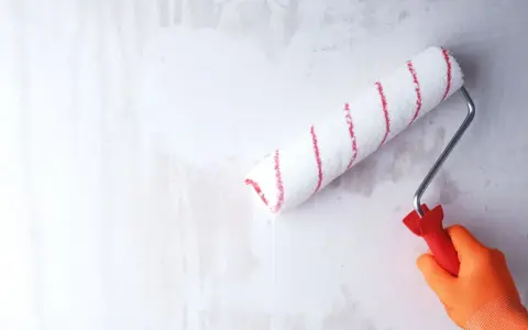 Ręka w pomarańczowej rękawicy gruntuje zeszklona ścianę wałkiem z czerwonymi paskami.