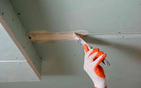 Ręka w rękawicy nakłada masę szpachlową na taśmę zbrojącą, przygotowując do klejenia płyt gk na grzebień.