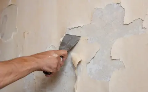 Dłoń zeskrobuje łuszczącą się farbę z gruntem ze ściany.