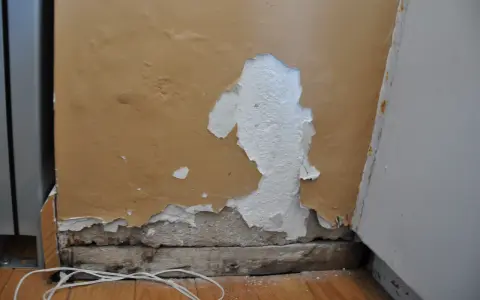 Zła gładź na ścianie, łuszcząca się farba odsłania tynk. Widoczne drewniane belki i biały kabel na podłodze.