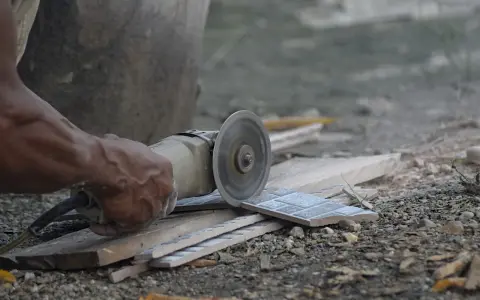 Ręka trzyma szlifierkę kątową, czym ciąć płytki gipsowe. W tle widać fragmenty płytek i gruzu.